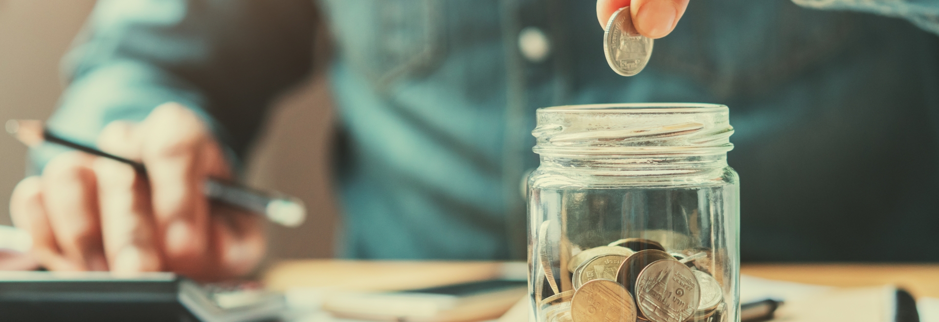 Man Holding Coin Above Jar While Using A Calculator In The Background